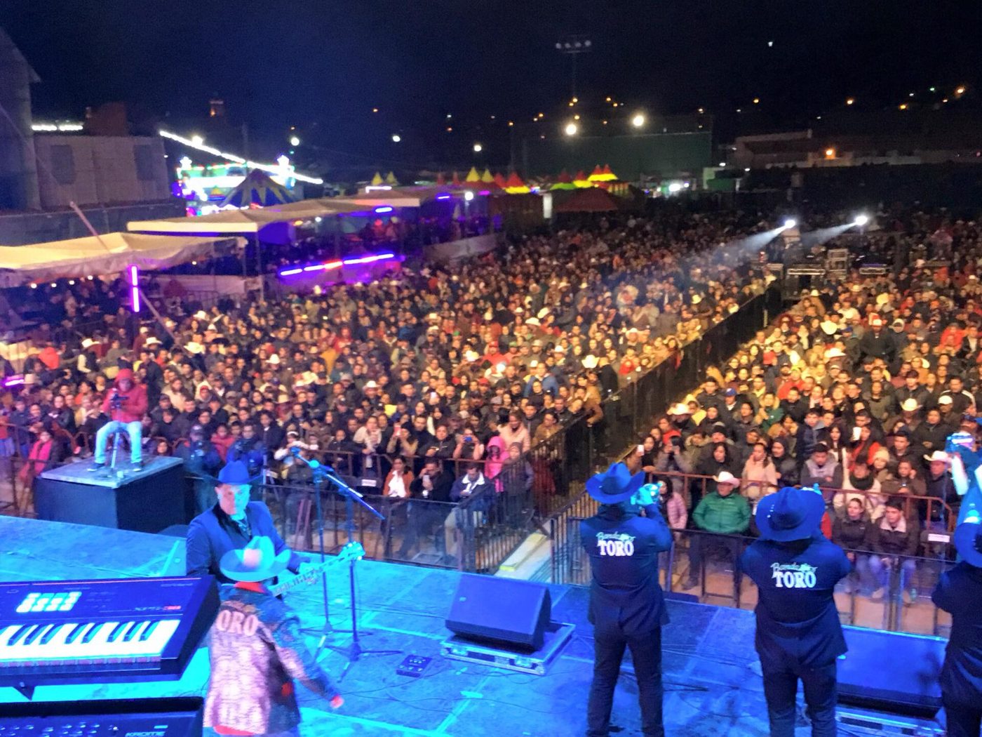 Escenario de noche con una banda tocando frente a una multitud, luces azules, tuba y percusión al frente.