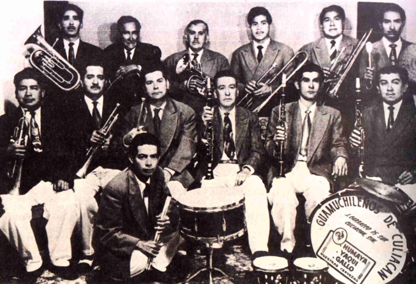 Fotografía antigua en blanco y negro de una banda sinaloense de dieciocho músicos con trompetas, trombones, tuba y tambora.