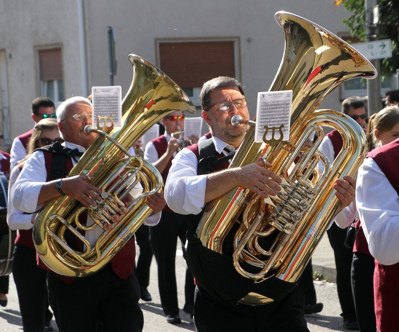 Dos músicos de una banda de viento alemana con tubas doradas, en chaleco negro y camisa blanca, desfilando por una calle.