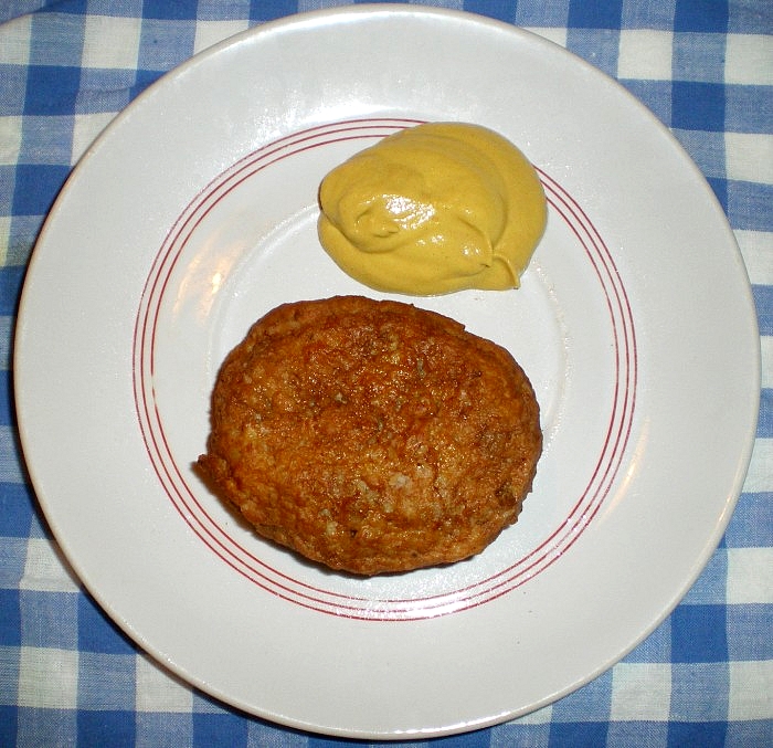 Una tortita de carne dorada en un plato blanco con filo rojo, junto a una cucharada de mostaza, sobre un mantel de cuadros azules y blancos.