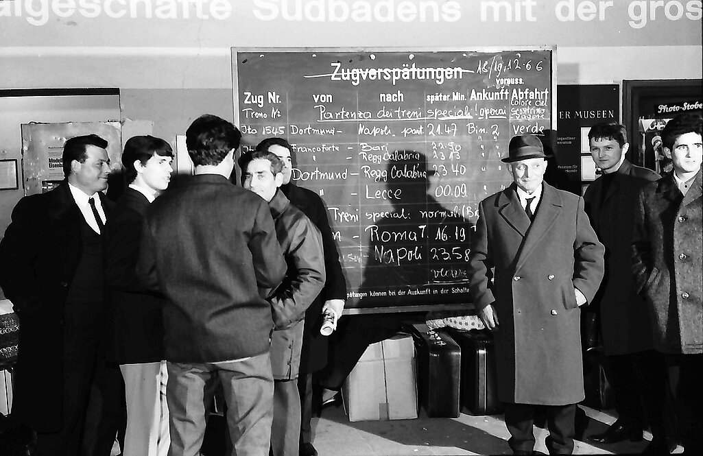Fotografía en blanco y negro de hombres con abrigo frente a un pizarrón de retrasos de trenes, con maletas en el piso. Estación central de Friburgo, diciembre de 1966.