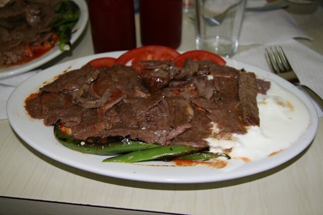 Un plato blanco con rebanadas delgadas de carne asada sobre pan, con yogur a un lado, jitomate y chile verde asado.
