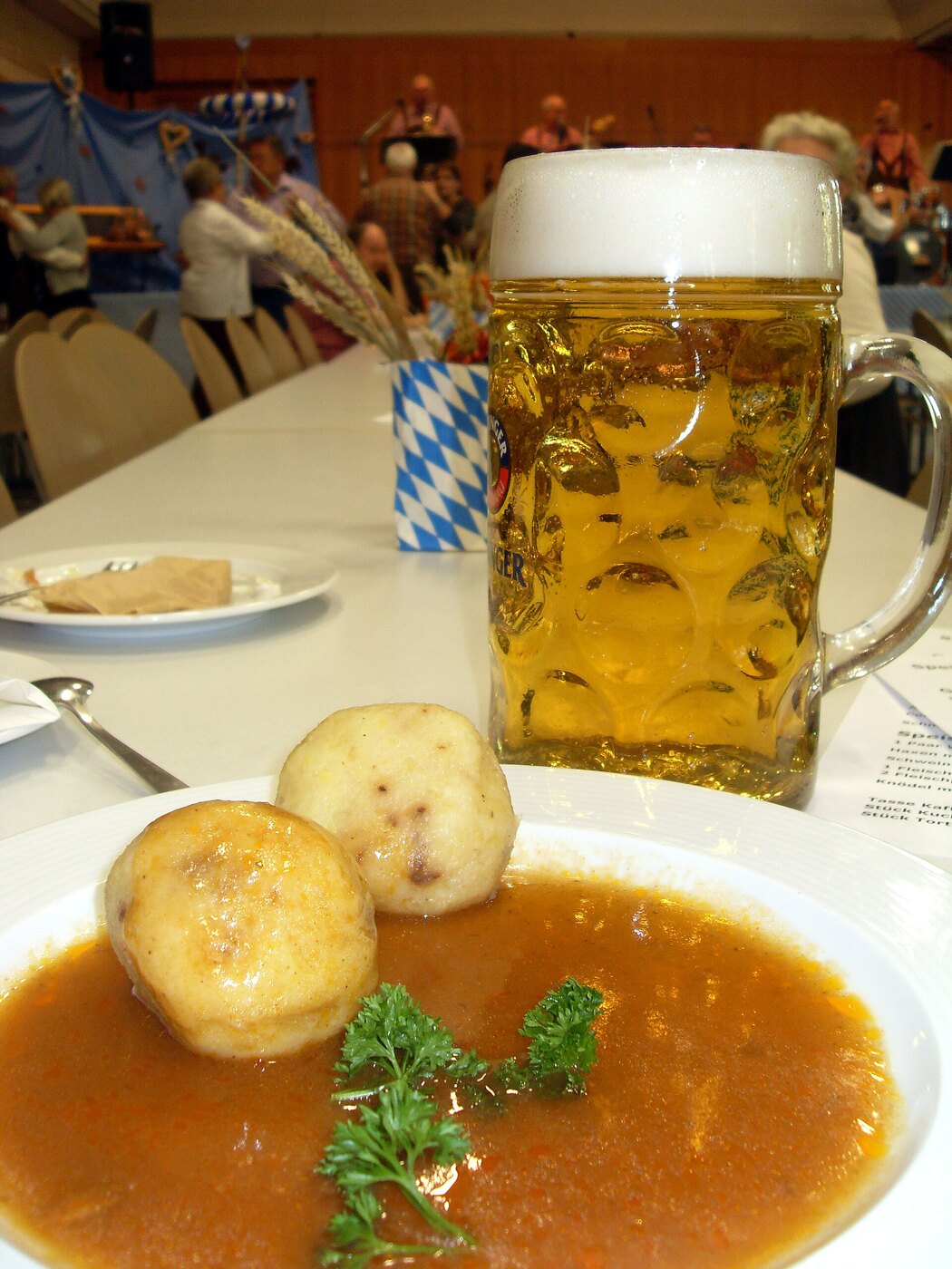 Dos bolas de papa en salsa oscura sobre un plato blanco, con un tarro de un litro de cerveza al fondo en un salón de fiesta.