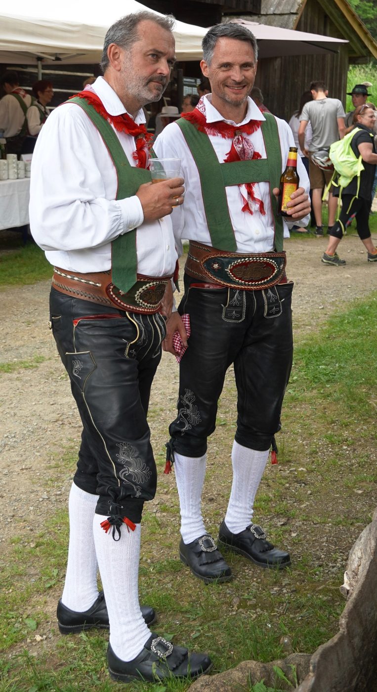 Cuatro hombres de un grupo de danza folclórica en Lederhosen y camisa blanca, de espaldas a un muro de piedra.