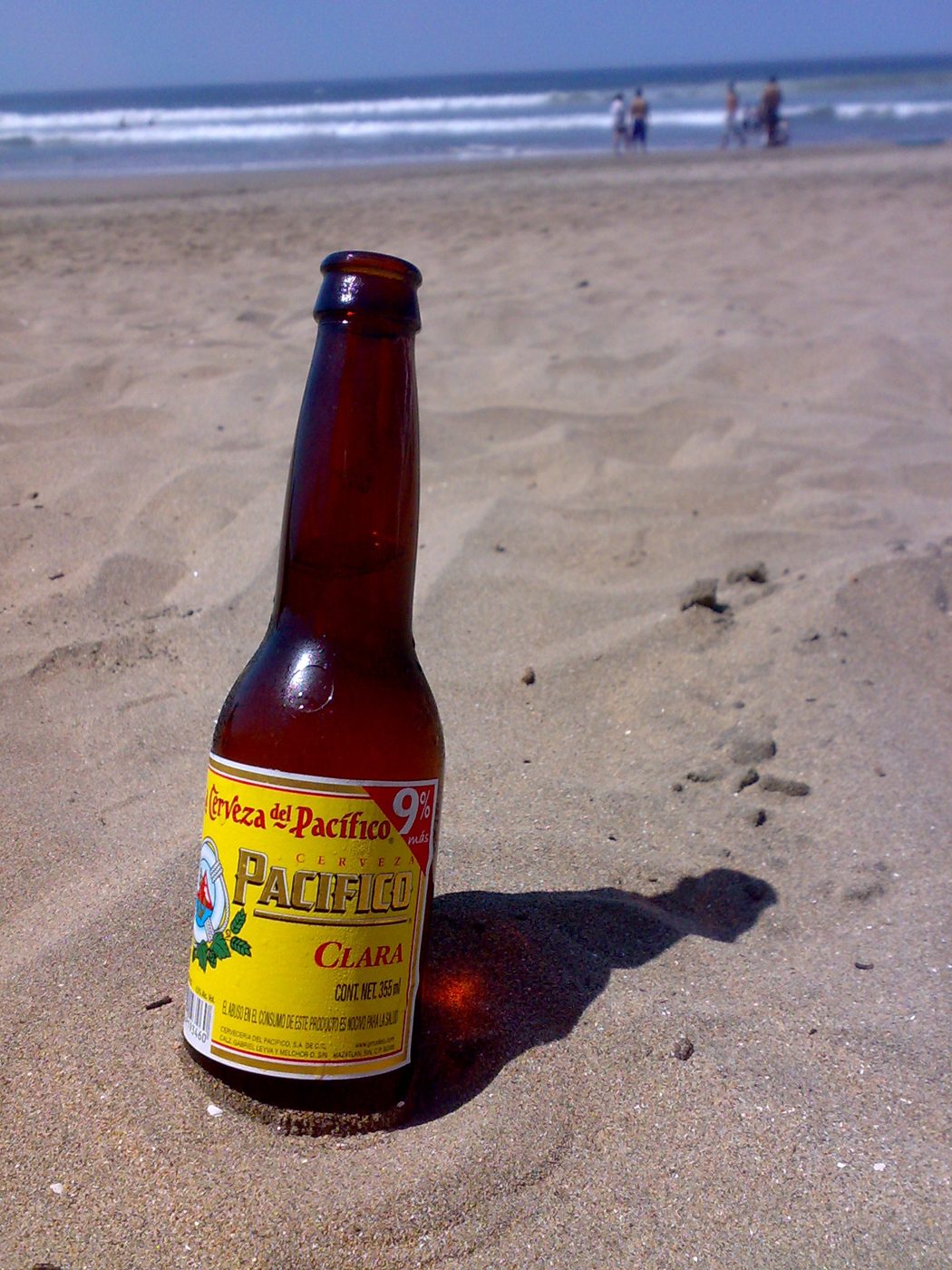 Botella de Pacífico Clara clavada en la arena de una playa, con el mar y bañistas al fondo.