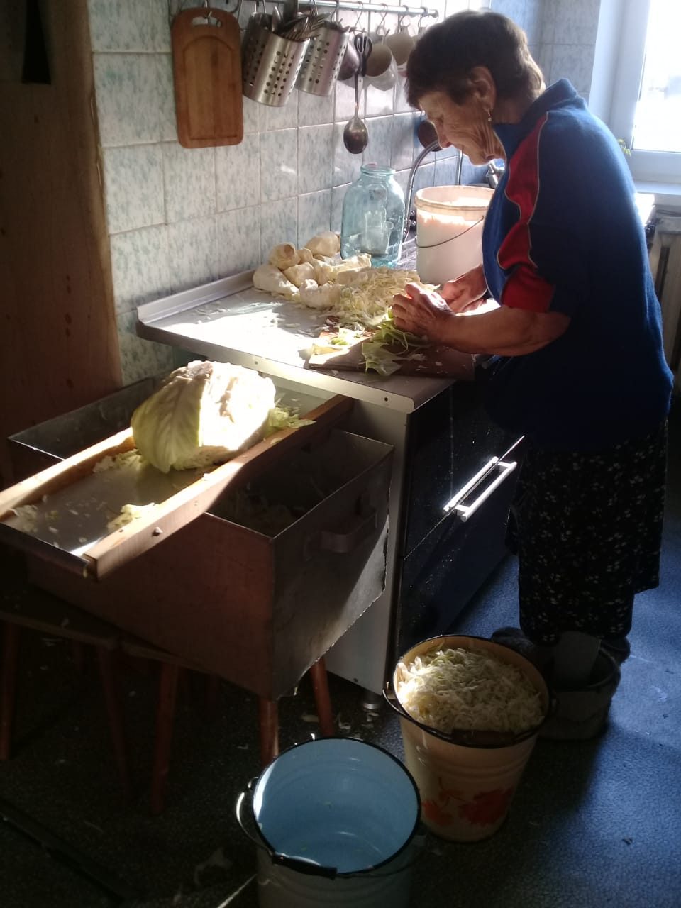 Una mujer en una cocina doméstica corta col a mano sobre una tabla, con cubetas de col picada en el piso.