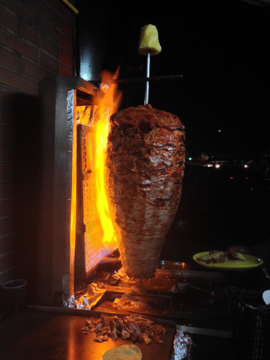 Un trompo de carne al pastor girando de noche frente a las llamas, con una piña encima y un plato amarillo al lado.